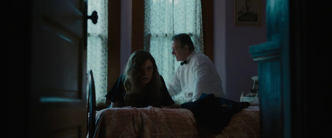 Live By Night (2016) movie still: medium — A dimly lit room with two windows draped in floral curtains behind a bed. A young woman and an older man…
