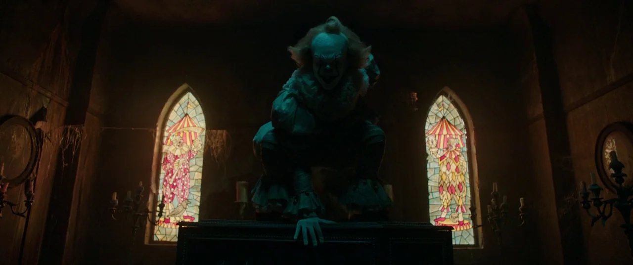 It (2017) movie still: fullbody — A dark, dilapidated room contains two stained glass windows depicting clowns. A clown crouches atop a dark…