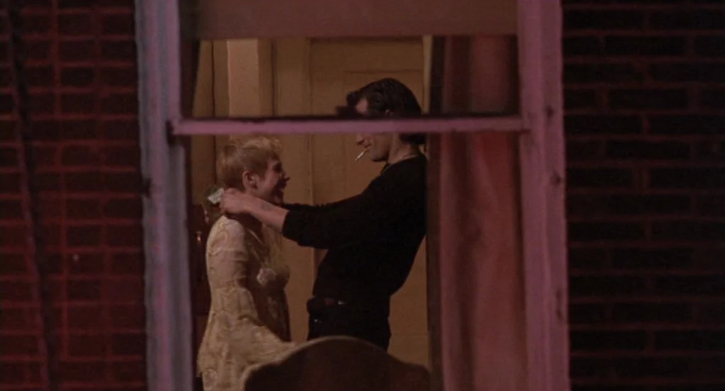 The Indian Runner (1991) movie still: medium — A dark brick wall frames a window view into a dimly lit residential space. A young man with a…