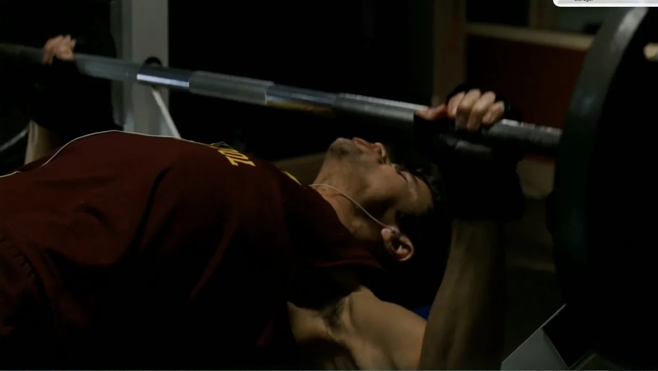 Chain Letter (2010) movie still: medium — A dimly lit indoor workout space features a weightlifting bench and heavy equipment. An athletic male…