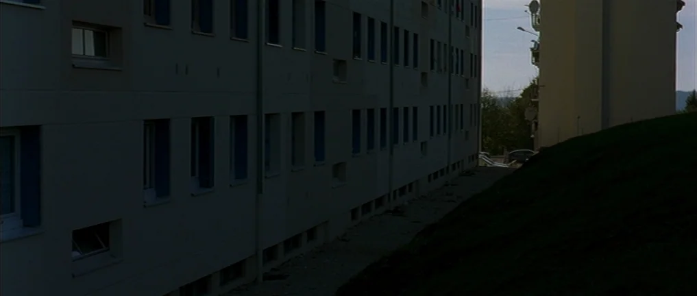 The Intruder (2004) movie still: establishing — A long, concrete residential apartment building sits beside a dark, grassy embankment. A narrow dirt…