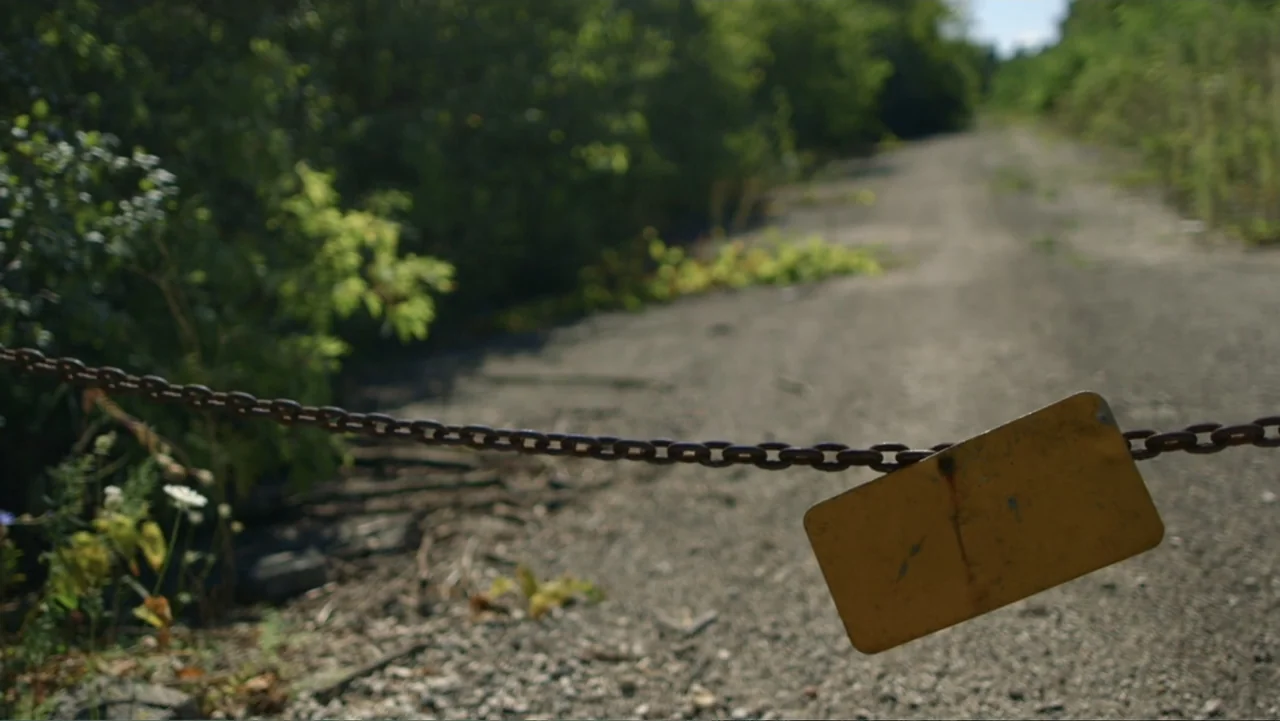 Beware the Slenderman (2016) movie still: wide — A rural dirt road is blocked by a rusted iron chain with a blank yellow sign. Lush green weeds and…