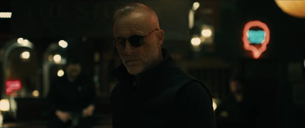 Guns Up (2025) movie still: medium — A dark, dimly lit bar environment with blurred neon signage and indistinct background figures. An older man in…