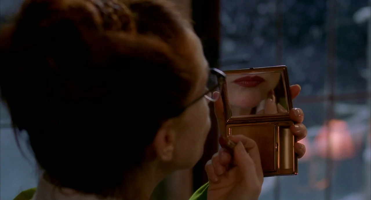 8 Women (2002) movie still: closeup — A woman holds a small golden compact mirror while indoors at night. She is carefully applying bright red…
