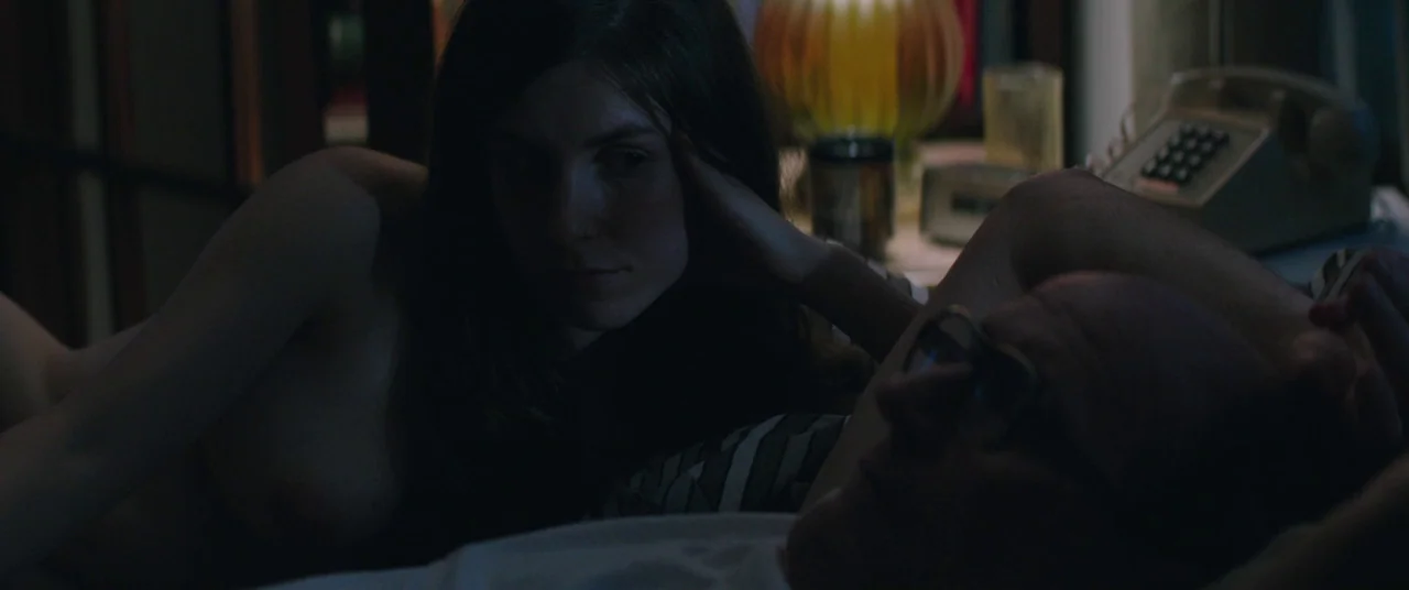 God’s Pocket (2014) movie still: medium — A dimly lit room at night features a bed with two people resting together. The woman rests her head on her…
