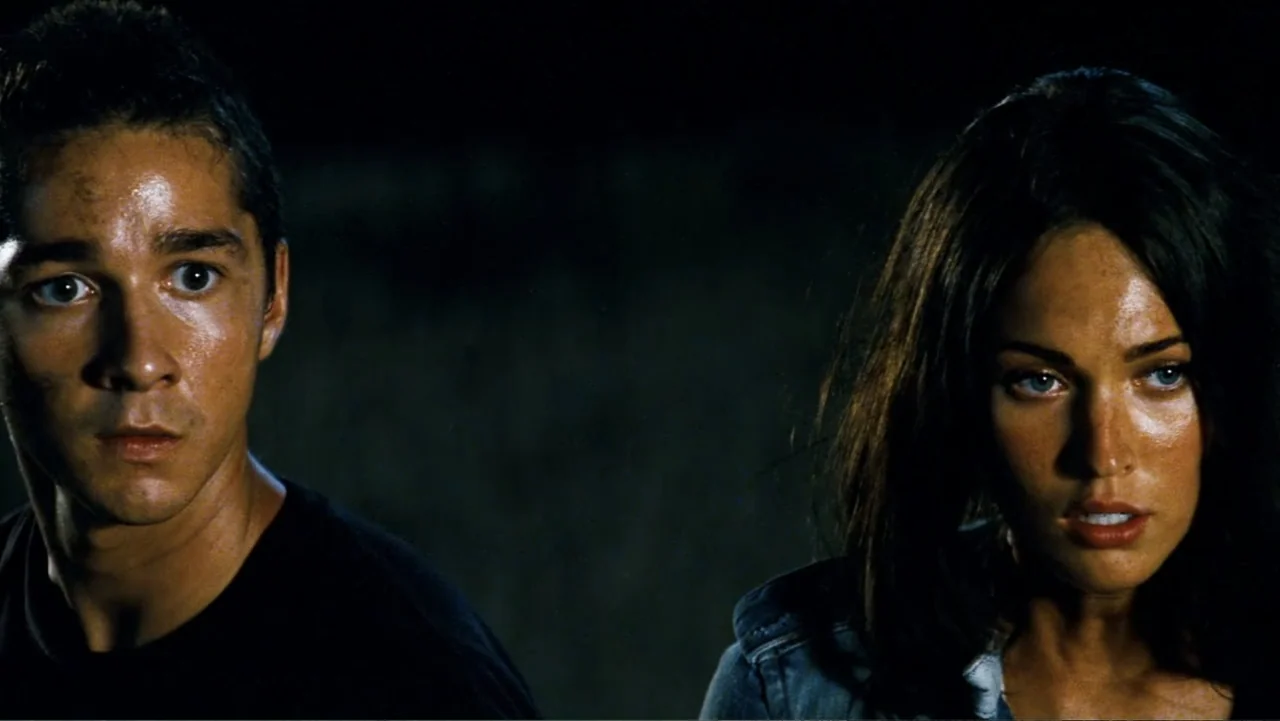 Transformers (2007) movie still: medium — Two individuals stand in a dark, nondescript night environment. The man and woman look intently toward the…