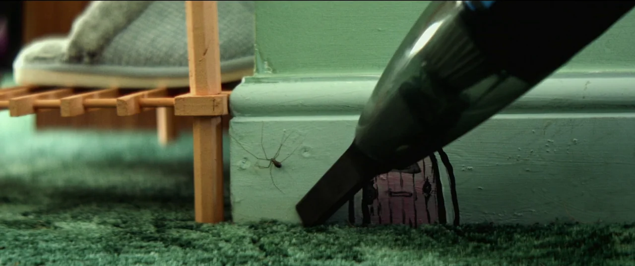 Scrapper (2023) movie still: closeup — A close-up of a carpeted floor showing a light green baseboard with a small drawing. A handheld vacuum nozzle…