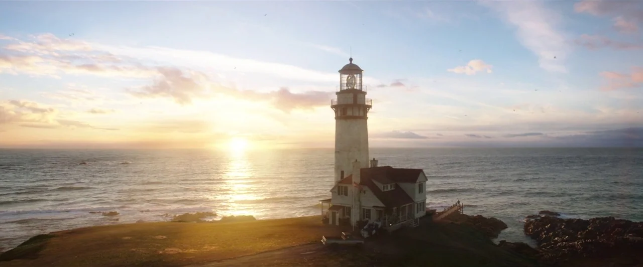 Aquaman (2018) movie still: establishing — A tall white lighthouse stands on a grassy cliff overlooking the ocean at sunset. The sky is filled with…