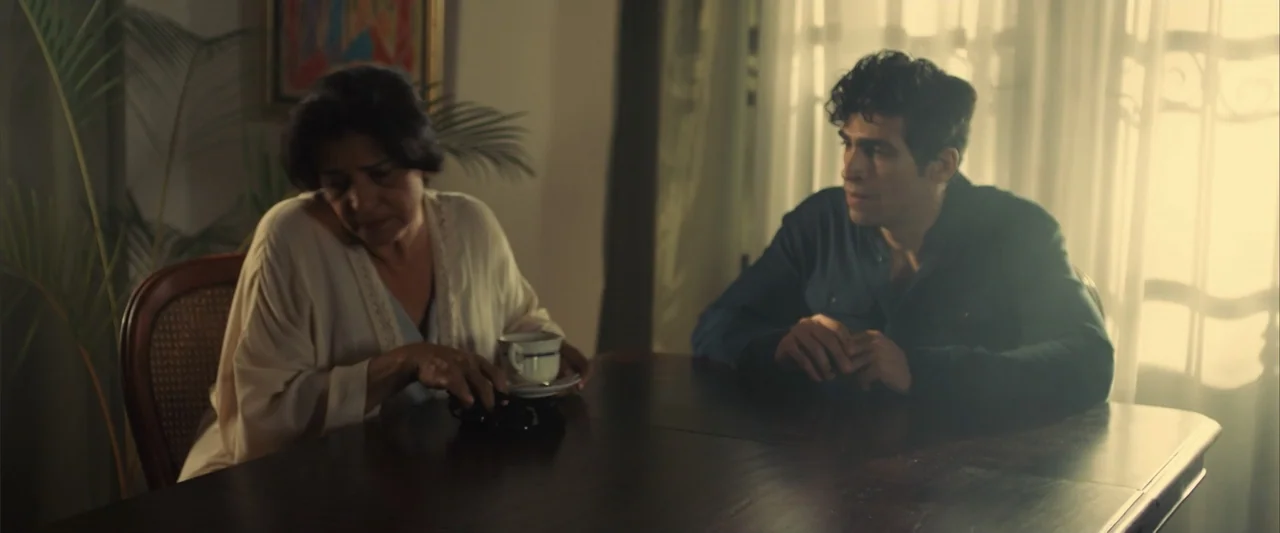 La Musiquita por Dentro (2019) movie still: medium — A quiet dining room with white curtains filtering daylight from a large window. A woman in a…