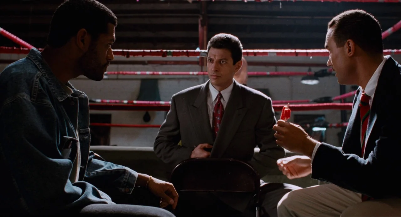 Deep Cover (1992) movie still: medium — Three men are seated in a dimly lit indoor boxing gym area. The men are engaged in a conversation while one…