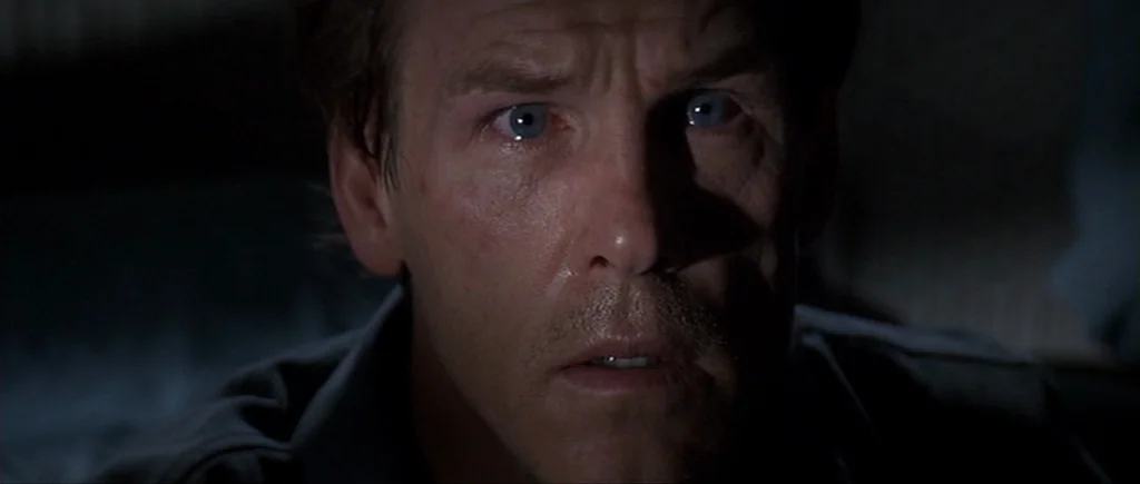 Cape Fear (1991) movie still: closeup — A dark, dimly lit environment obscures all background details behind the subject. A man stares forward while…