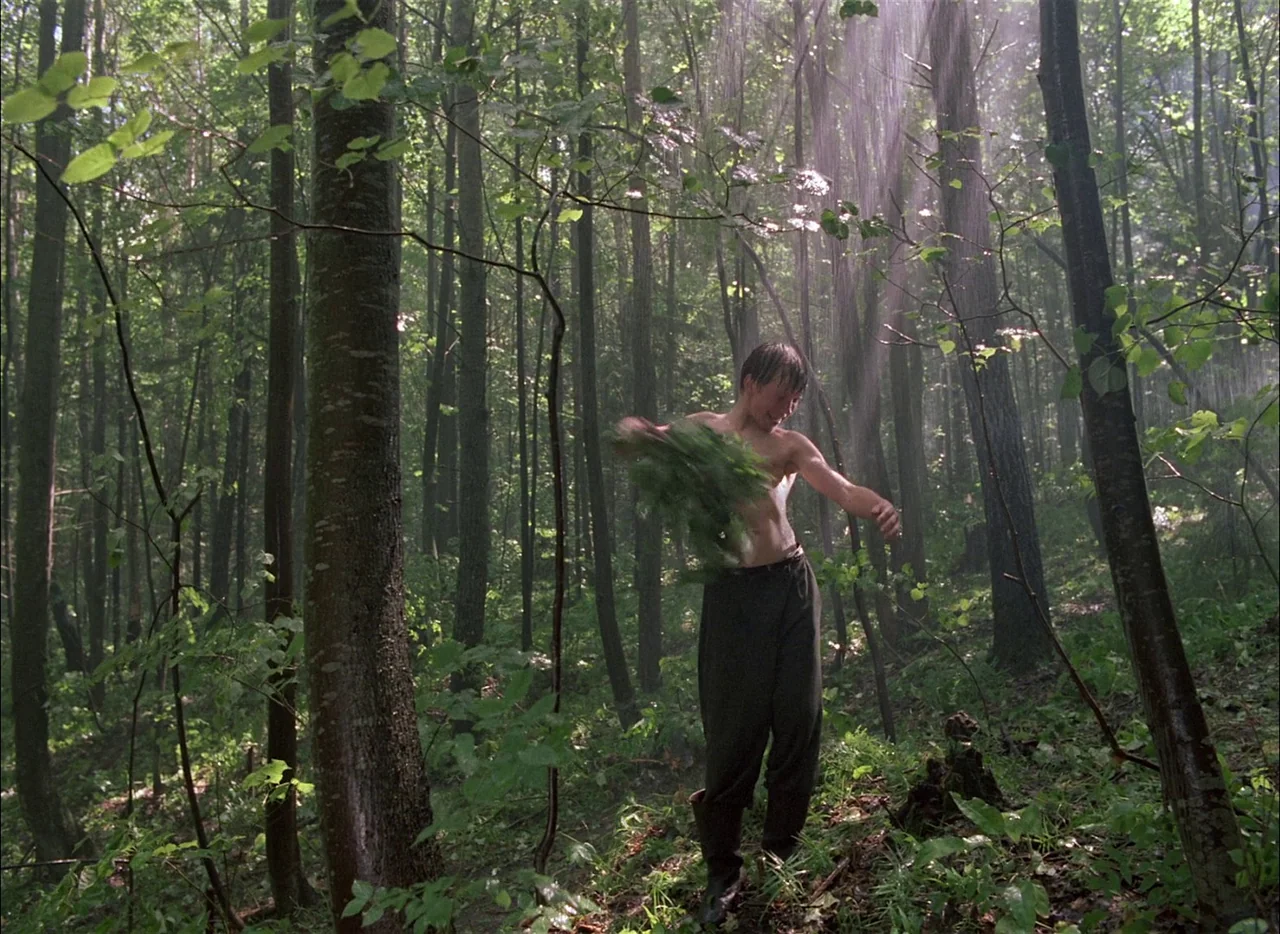 Come and See (1985) movie still: fullbody — A dense woodland with tall slender trees and lush green undergrowth. A bare-chested boy holds a large…