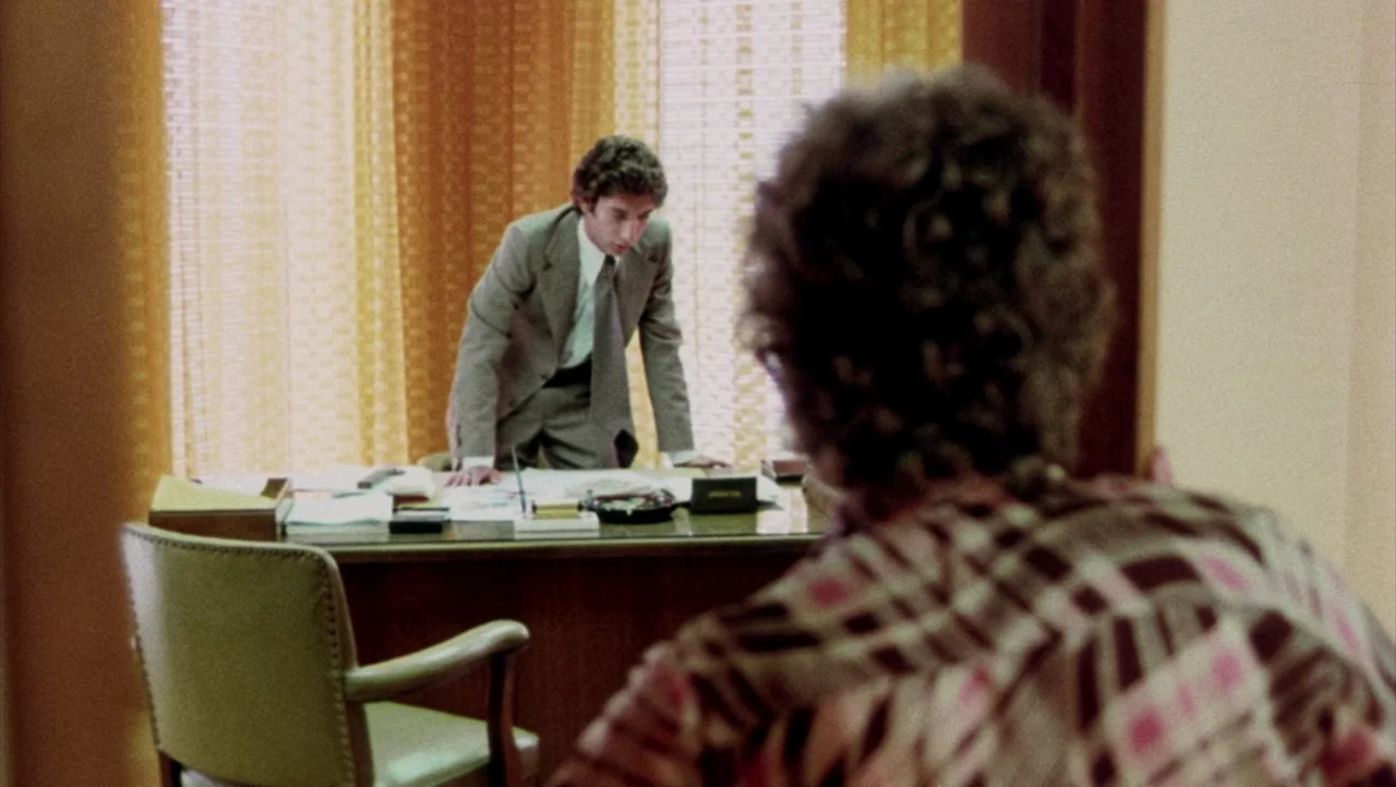 Shivers (1975) movie still: medium — A 1970s workspace with a wooden desk and heavy yellow curtains. A man in a suit leans over the desk speaking to…
