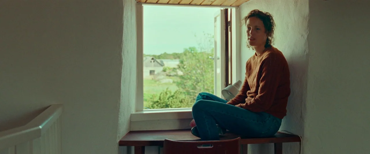 Bergman Island (2021) movie still: medium — A rustic interior features a wooden desk integrated into a deep window frame. A woman sits on the desk…