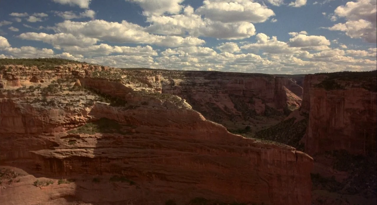Koyaanasquatsi (1982) movie still: establishing — A vast red sandstone canyon stretches across the horizon under a bright blue sky. Numerous white…