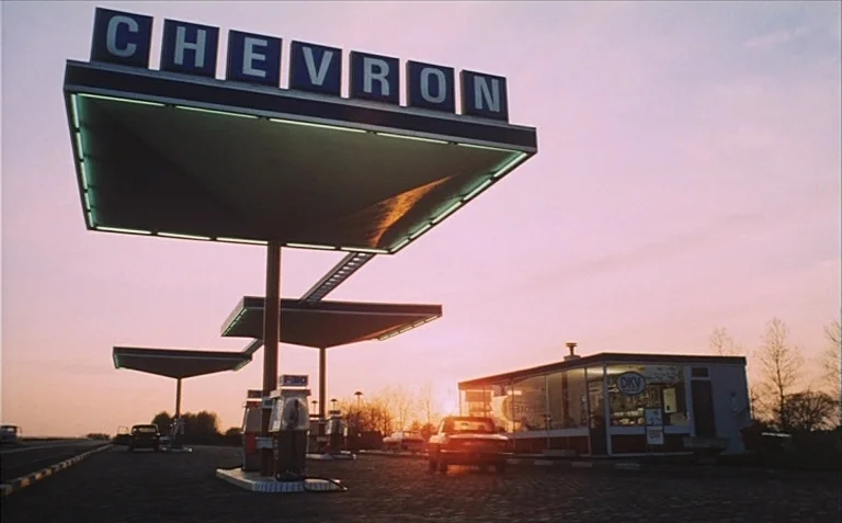 Business is Business (1971) movie still: wide — A Chevron gas station with distinctive geometric canopy structures sits under a pink sunset sky. The…
