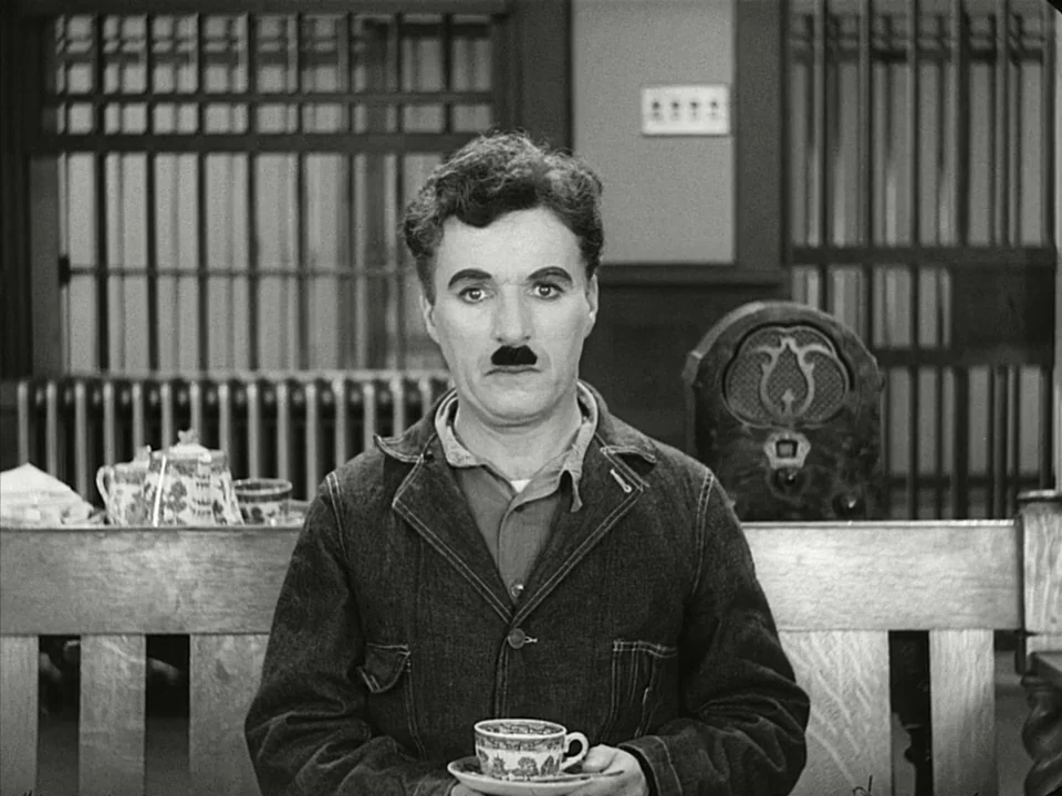 Modern Times (1936) movie still: medium — A spare interior space features a wooden bench, a radiator, and a vintage radio. A man in a dark jacket…