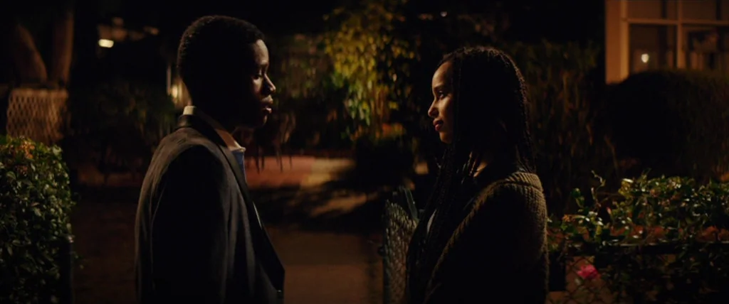 Dope (2015) movie still: medium — A residential sidewalk at night with bushes flanking a paved path. A man and woman stand face to face in a quiet…