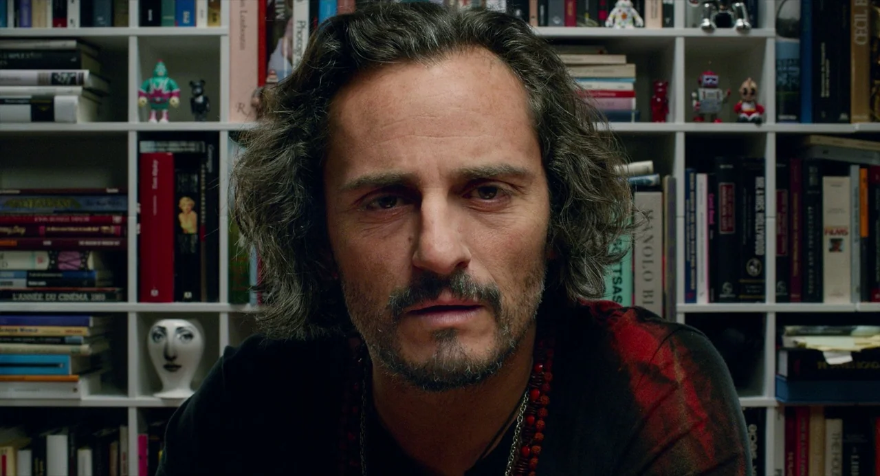 Pain and Glory (2019) movie still: closeup — A white bookshelf packed with colorful books and small collectible figurines forms the background. A…