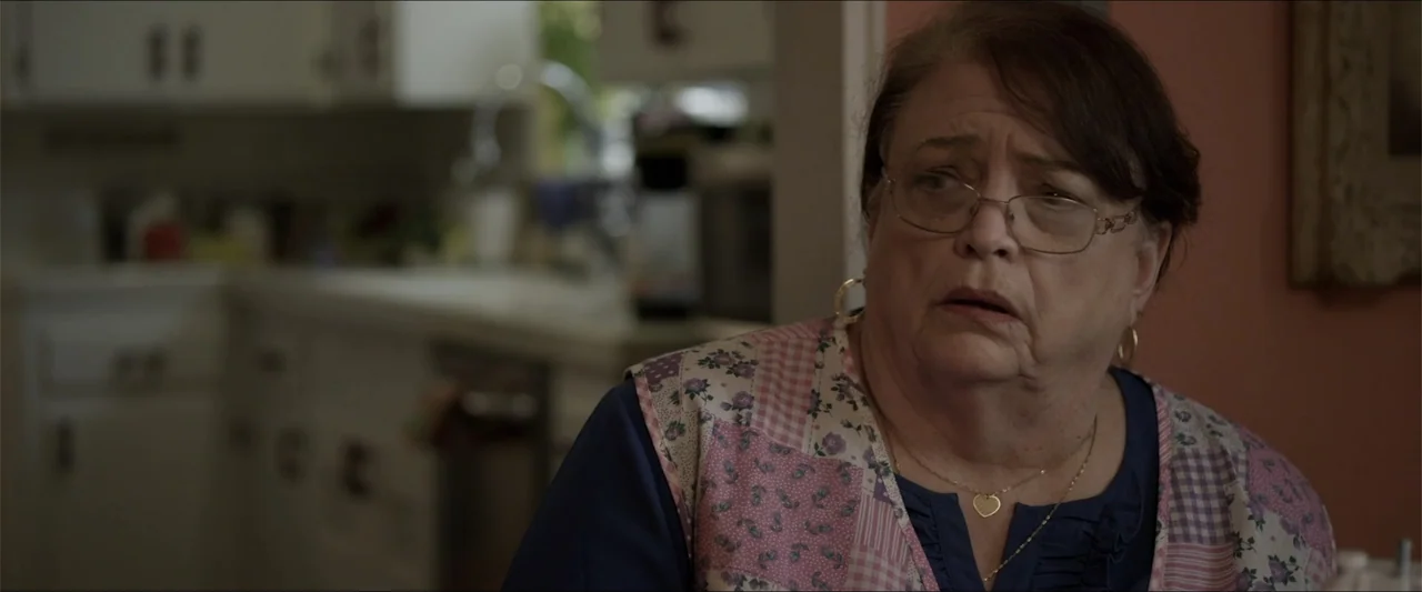 Heavenly Deposit (2019) movie still: medium — A residential dining room area is visible with an out-of-focus kitchen background. An older woman with…