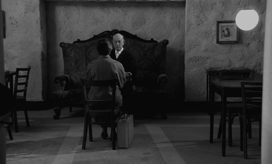 The Man From London (2007) movie still: wide — A sparse interior with textured walls and tables, featuring a classic couch and wooden chairs. A…
