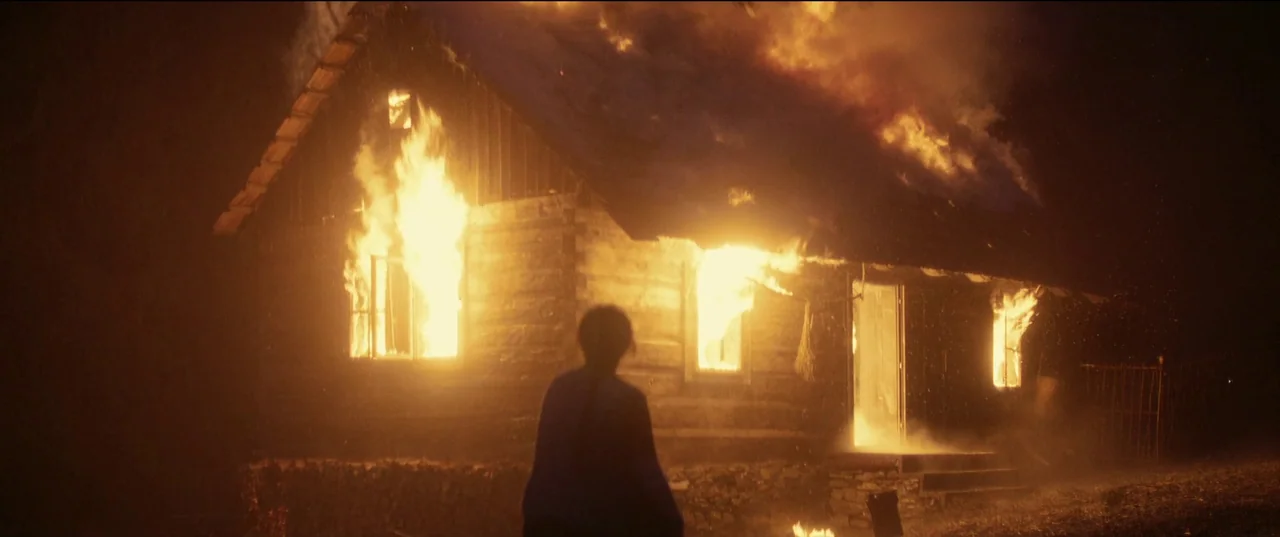 Nightsiren (2022) movie still: wide — A dark wooden log cabin engulfed in bright orange flames at night. A lone figure stands in the foreground…