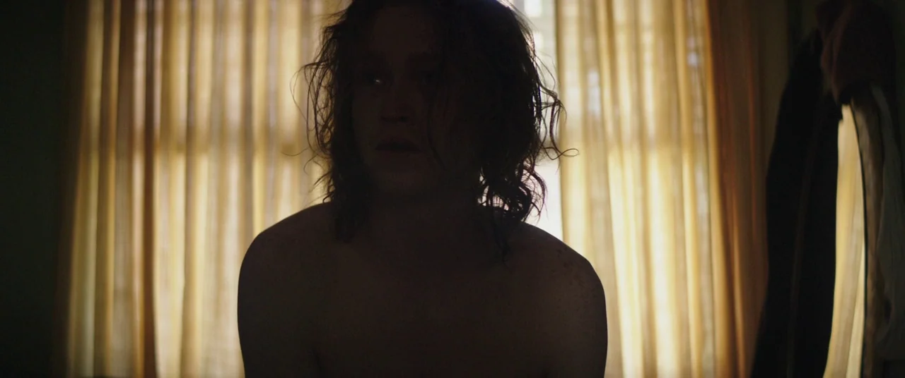 God’s Pocket (2014) movie still: medium — A shirtless person sits indoors against a window covered by thin yellow curtains. The bright light behind…