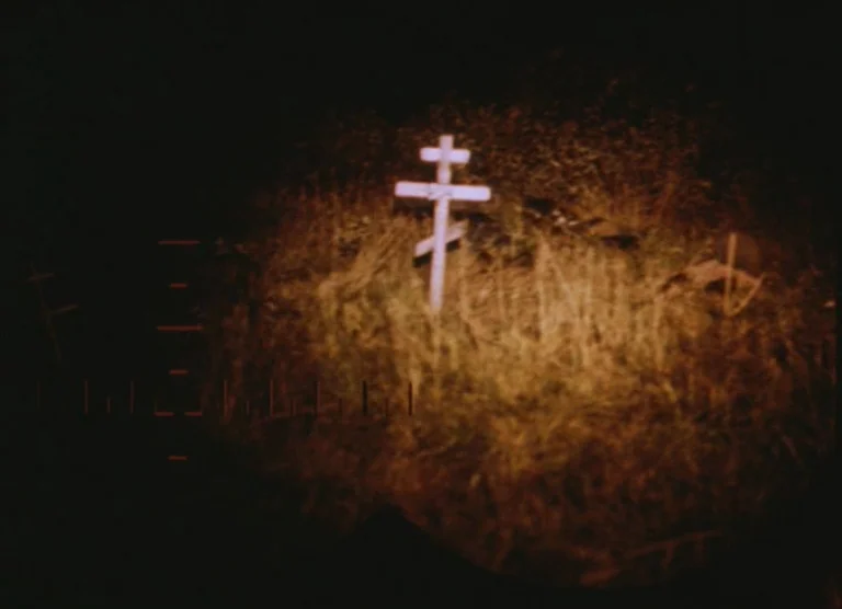 Russian Symphony (1994) movie still: wide — A lone white Orthodox cross stands in a grassy field at night. A bright spotlight beam targets the cross…