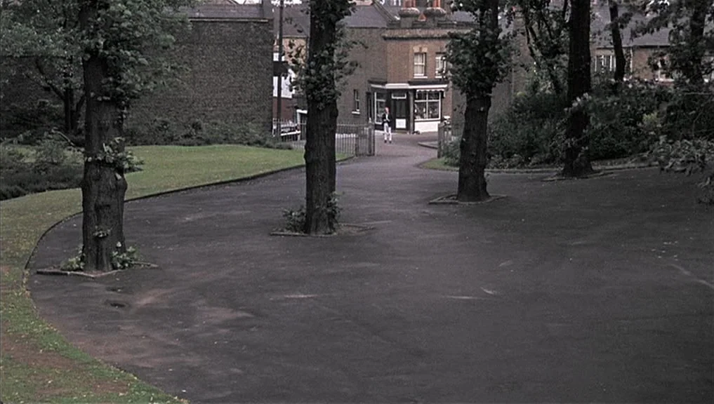 Blow-Up (1966) movie still: wide — A paved path winds through a park bordered by mature trees and grass. Brick buildings are visible in the…