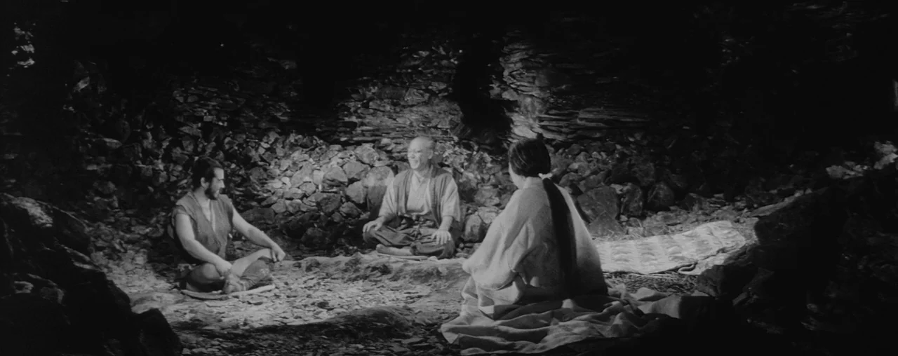 The Hidden Fortress (1958) movie still: wide — A rough, uneven cave interior with dark stone walls and a dirt floor. Three people sit together in…