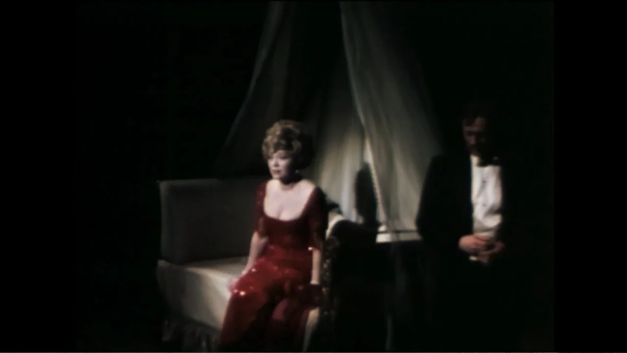 Six by Sondheim (2013) movie still: medium — A dimly lit bedroom features a bed with a white canopy and dark surroundings. A woman in a red dress…