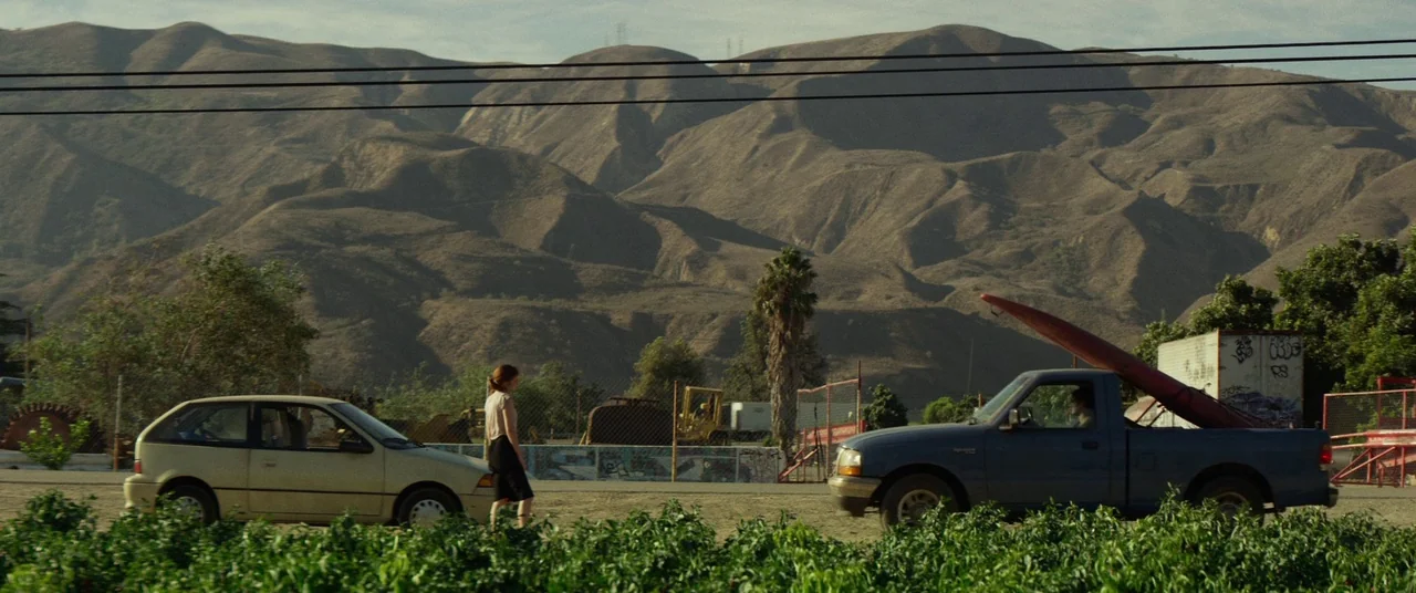 The Circle (2017) movie still: wide — A wide shot shows a dusty field beneath rolling brown mountains under bright sunlight. A woman walks past a…