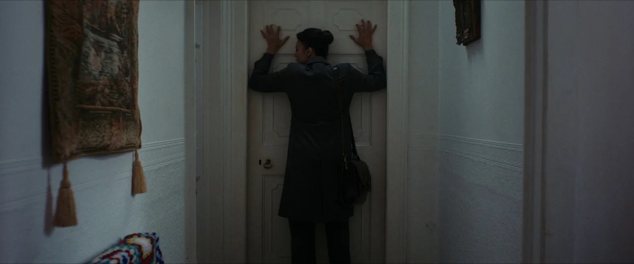 Fréwaka (2024) movie still: medium — A narrow domestic hallway with white walls features a tapestry and a wooden door. A woman stands in front of…