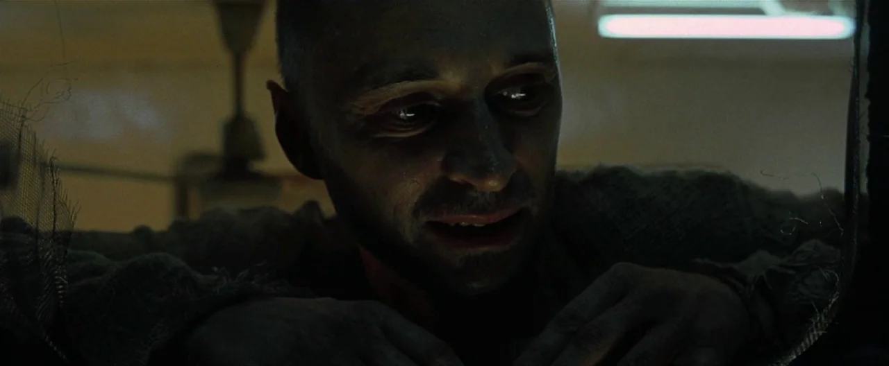 The Beach (2000) movie still: closeup — A dimly lit indoor space with a stark green-tinted fluorescent glow. A man with a shaved head and stubble…