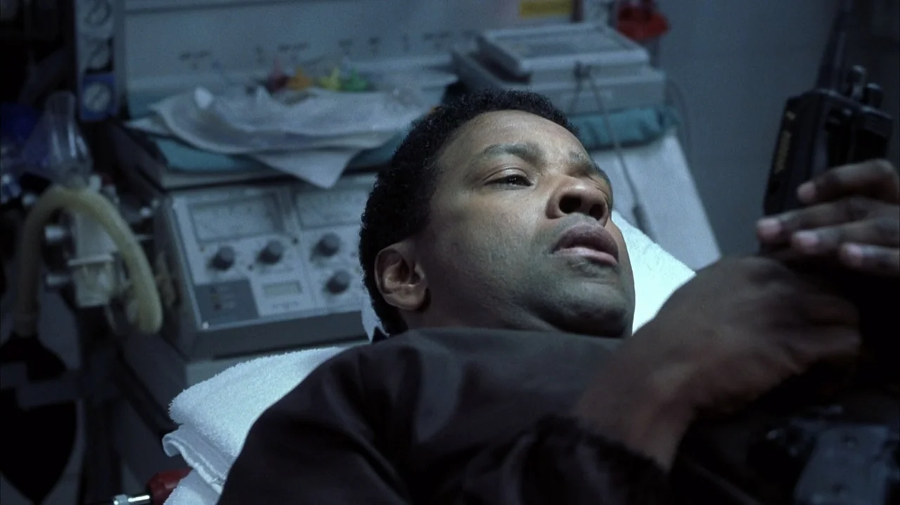 John Q (2002) movie still: closeup — A hospital room contains medical diagnostic machinery in the blurred background. A man lies on a white medical…