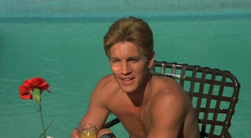 The Coca-Cola Kid (1985) movie still: medium — A man sits shirtless in a patio chair beside a bright turquoise swimming pool. A single red carnation…