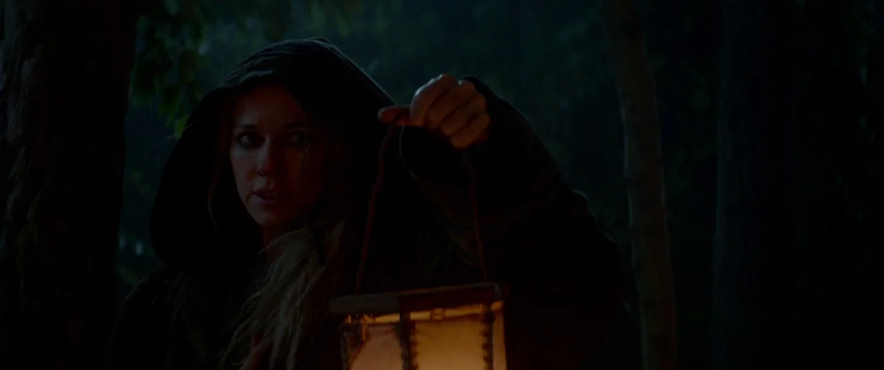 Ophelia (2018) movie still: medium — A dense, dark forest at night provides the backdrop for a hooded woman walking. She holds a small,…