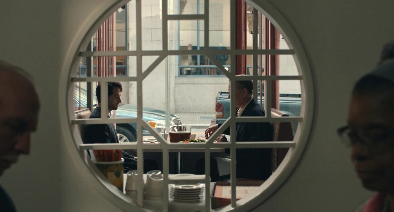 A Beautiful Day In The Neighborhood movie still: medium — A diner booth is framed through a circular wooden cutout with a grid pattern. Two men sit…