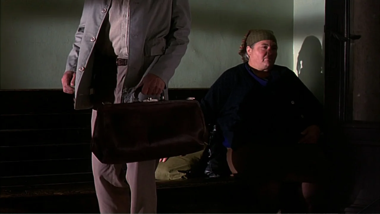 Citizen X (1995) movie still: medium — A dimly lit interior space features a wooden bench against a painted wall. A man holding a leather bag stands…