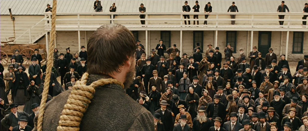 True Grit (2010) movie still: medium — A crowd of people in 19th-century attire stands in a large outdoor town plaza. A man with a thick rope noose…