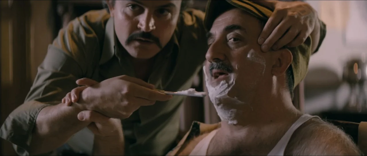 A Farewell to Fools (2013) movie still: medium — A dimly lit room with wooden textures serves as a private grooming area. One man holds a razor to…