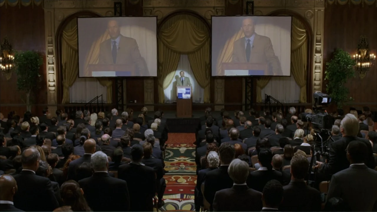 Rumor Has It... (2005) movie still: wide — An ornate interior event space features a central stage with a lectern and projectors. A man presents to…