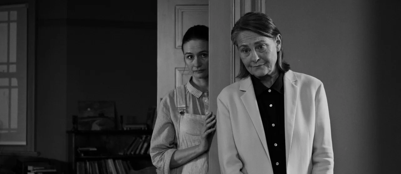 The Party (2017) movie still: medium — Two women stand in a doorway within a quiet, dimly lit domestic space. The older woman is positioned in front…