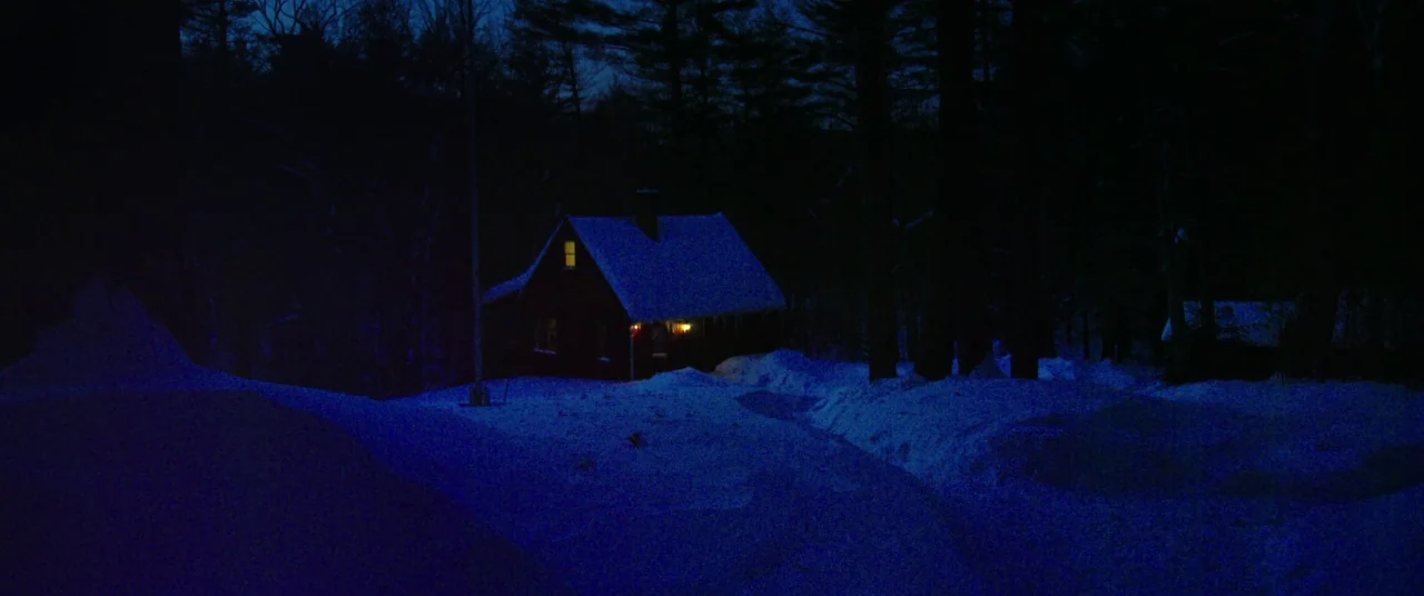 The Mind’s Eye (2015) movie still: wide — A dark wooden house sits nestled within a dense cluster of towering pine trees. Thick snow blankets the…