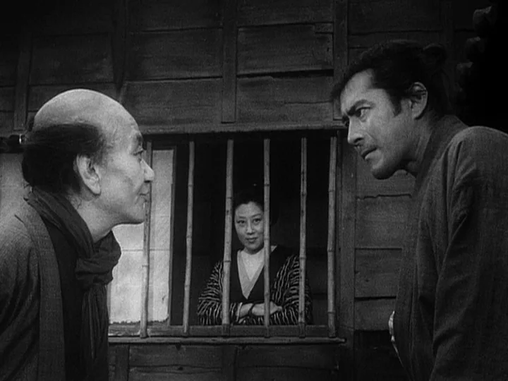 The Lower Depths (1957) movie still: medium — A wooden exterior of a building features a small window with vertical bamboo bars. Two men stand in…