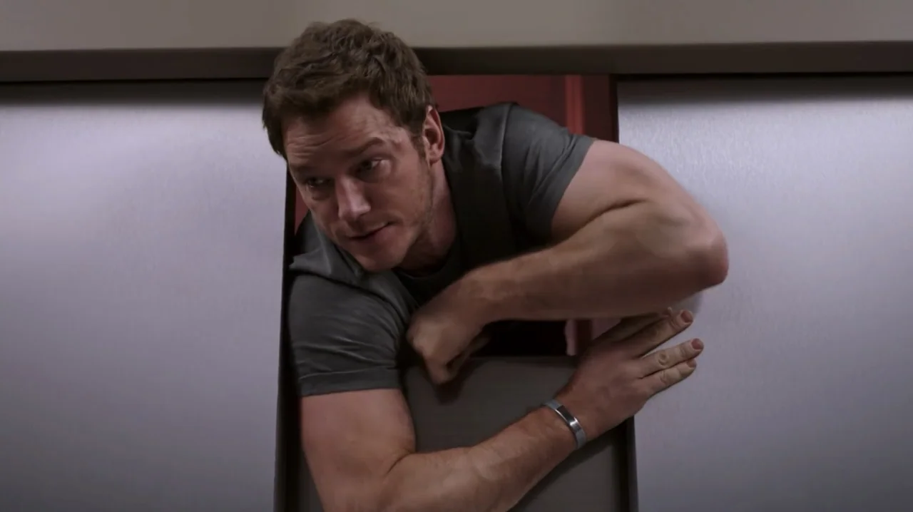 Passengers (2016) movie still: medium — A man peers through a narrow vertical gap between two metallic sliding elevator doors. The man has his hands…