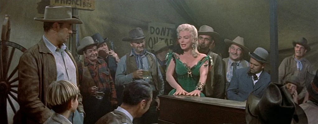 River of No Return (1954) movie still: wide — A crowded tent saloon interior features canvas walls and rustic wood furniture. A blonde woman in a…