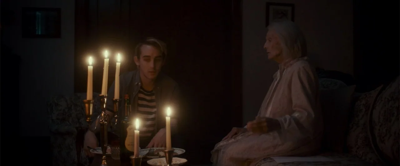 Jump, Darling (2022) movie still: medium — A dimly lit interior room at night features a table between two people. A young man and an elderly woman…