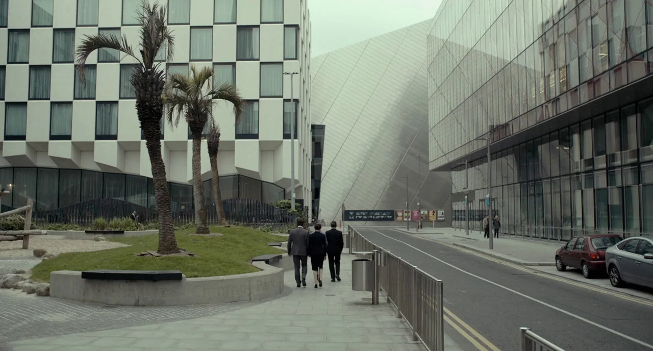 The Lobster (2015) movie still: wide — A modern city street bordered by large glass-fronted buildings and a landscaped patch. Three professionals in…