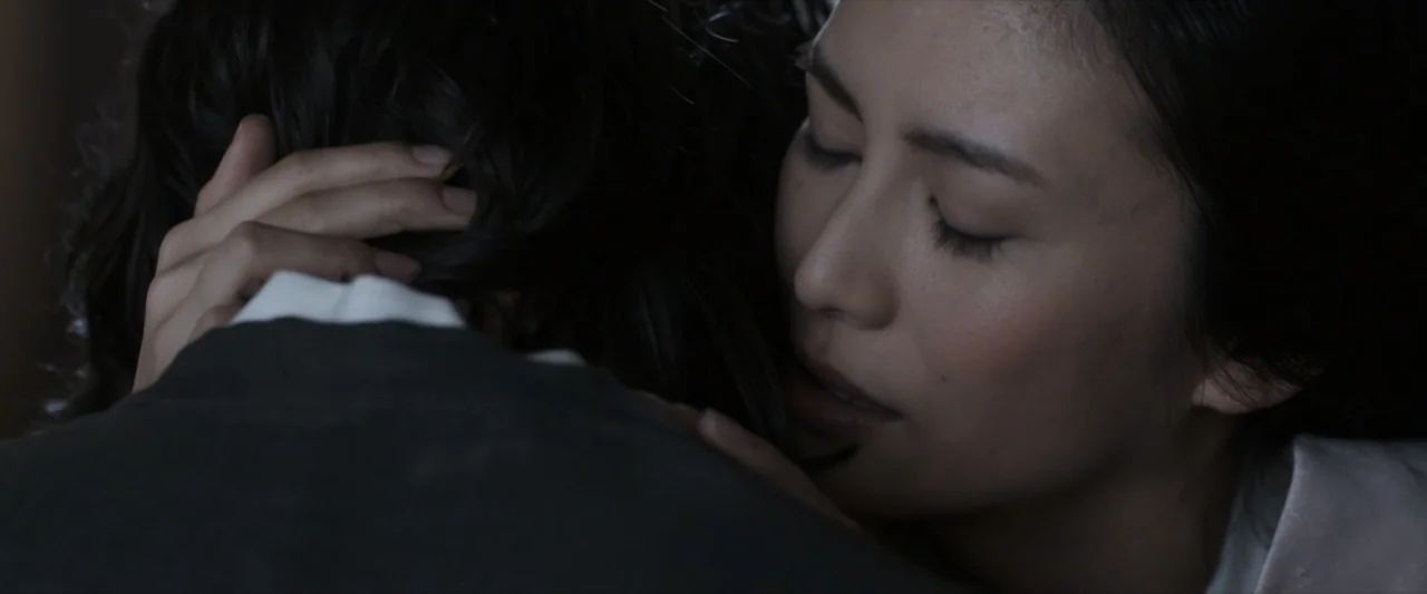 47 Ronin (2013) movie still: closeup — A dimly lit interior space with minimal architectural detail visible. One woman embraces another person while…